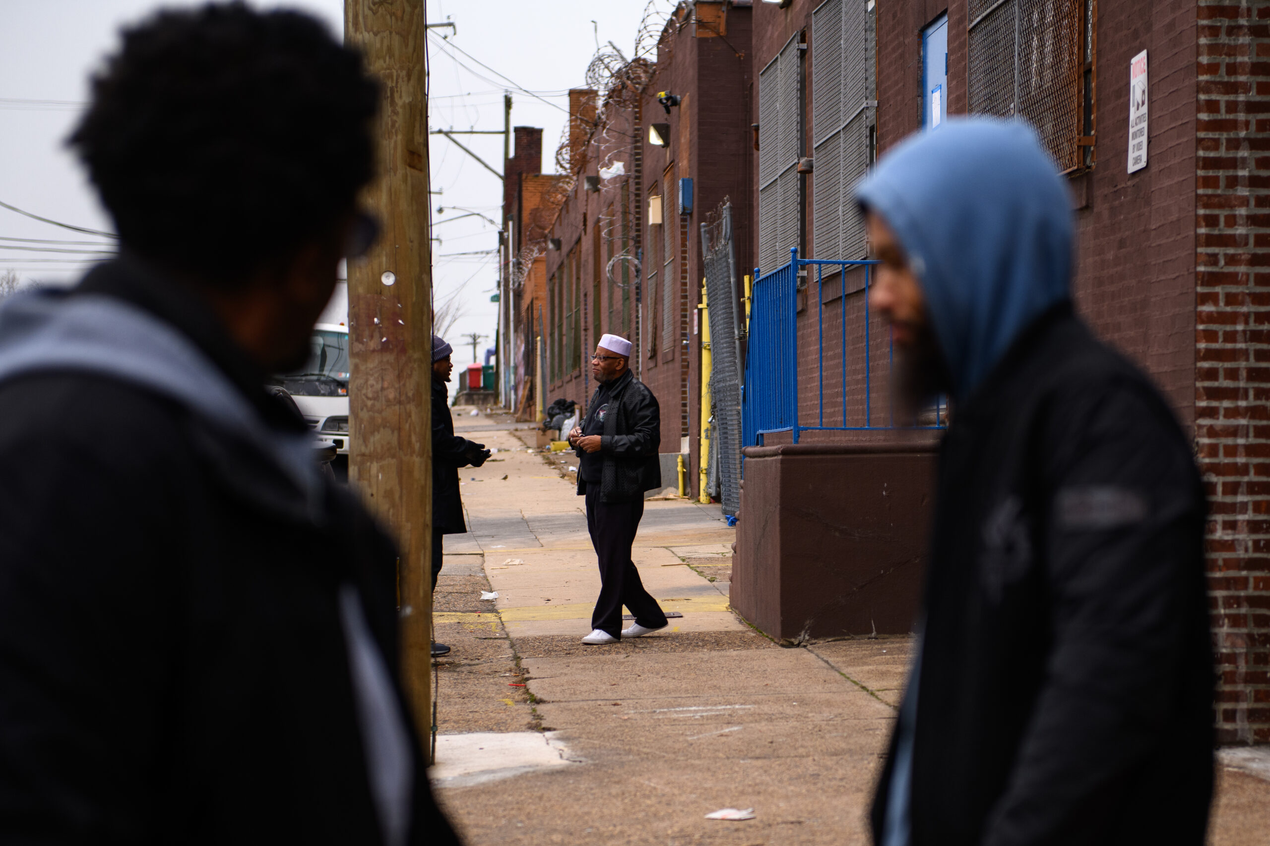 A photograph with two Black men in the foreground and another in the background, wearing a white kufi hat and having a conversation with a fourth man